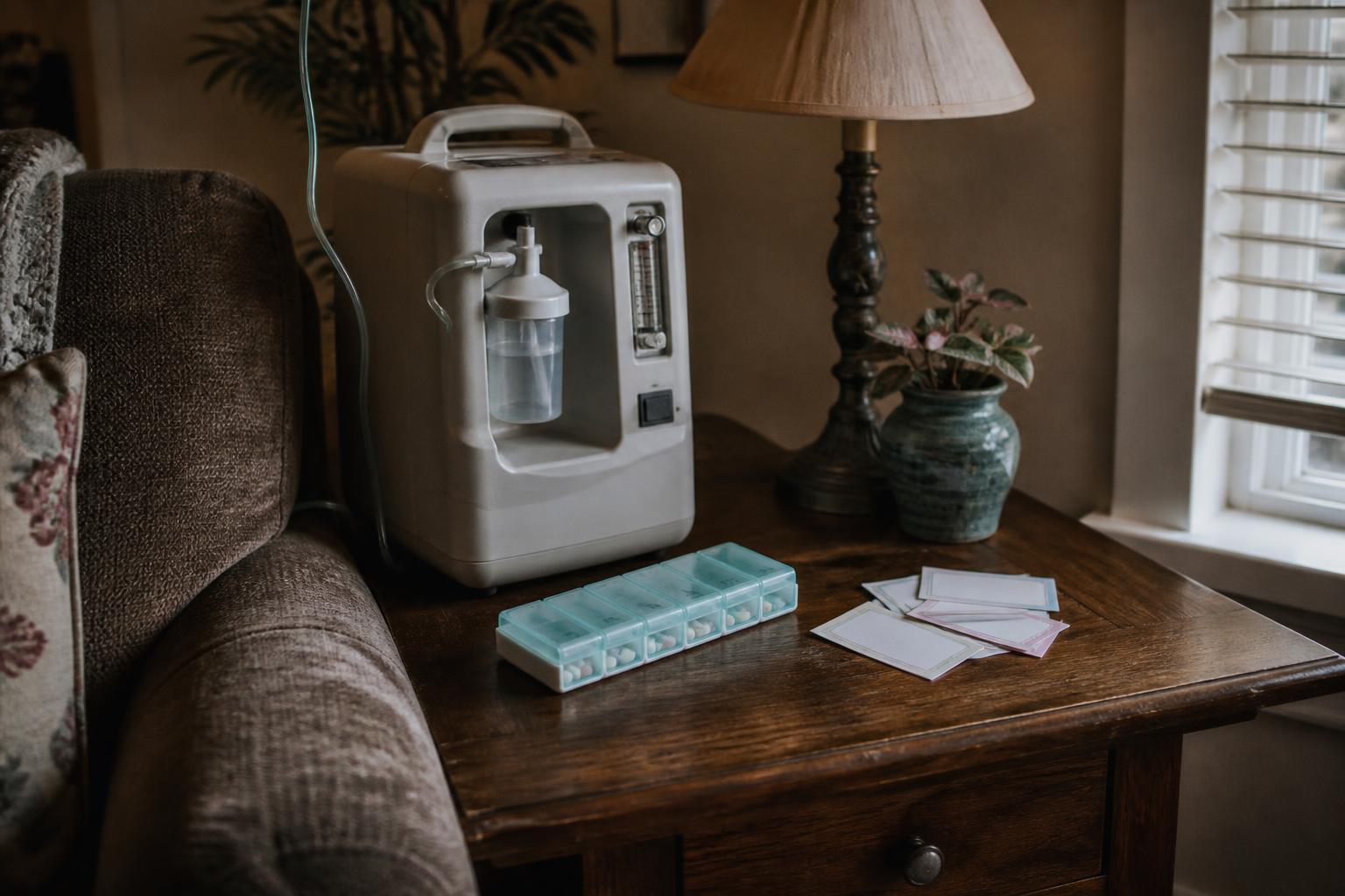An oxygen concentrator, pill organizer, lamp, and papers sit beside a couch