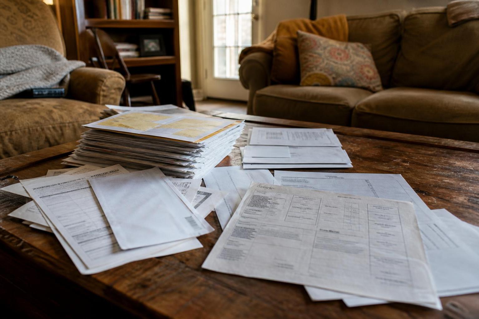 Scattered papers and stacked envelopes spread across a wooden coffee table in a living room